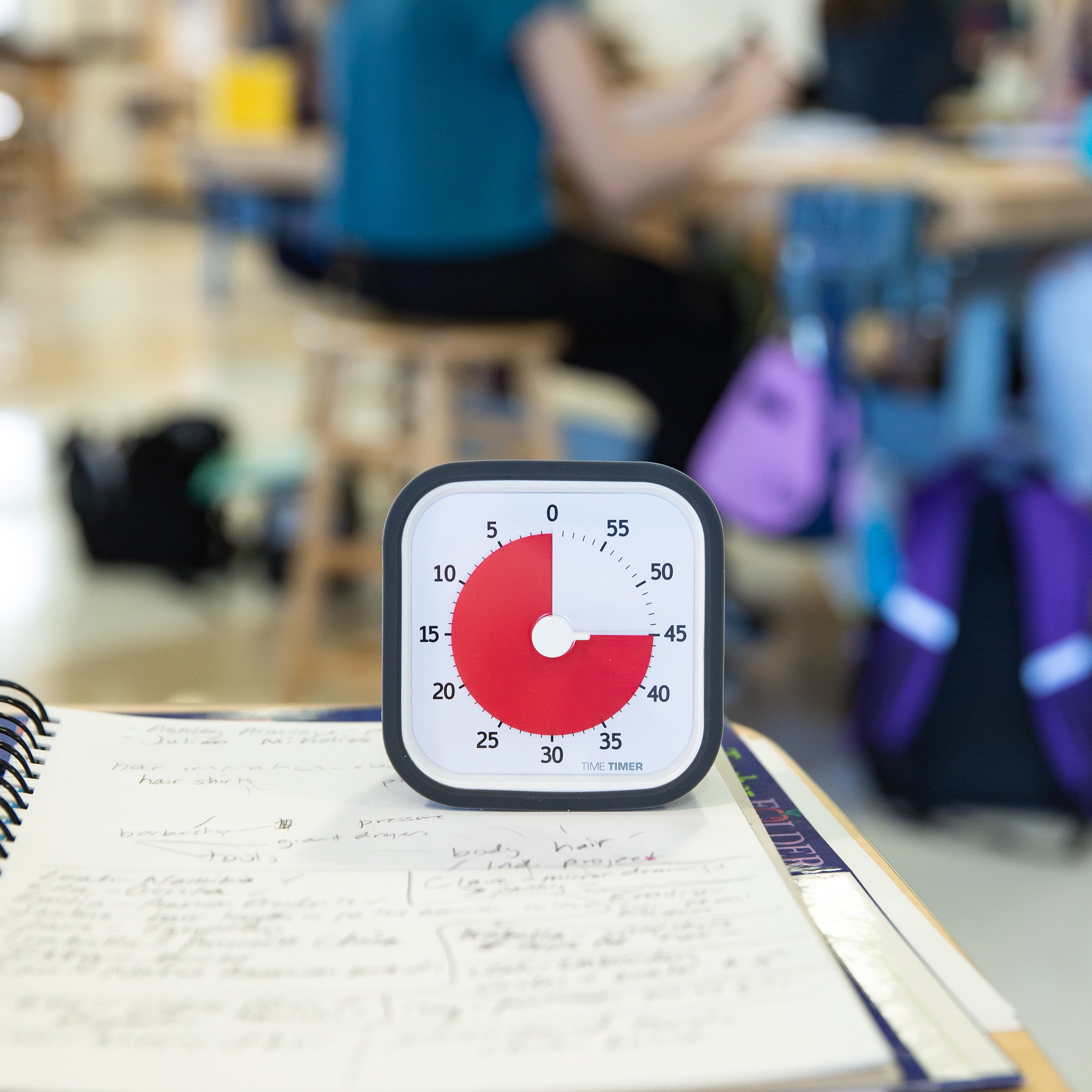 The Time Timer MOD 60 Minute visual timer is shown sitting on a notebook in a classroom setting. The red disk is set to 45 minutes and is shown as a three-quarters pie shape.