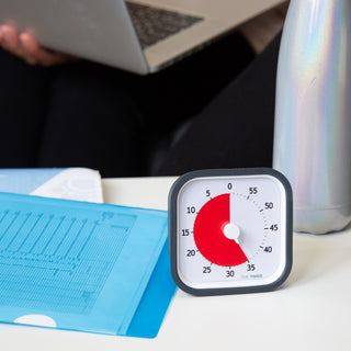 The Time Timer MOD 60 Minute visual timer is shown on a desk with spreadsheets and a water bottle. There is someone in the background on a computer. The red disk is set for 35 minutes.