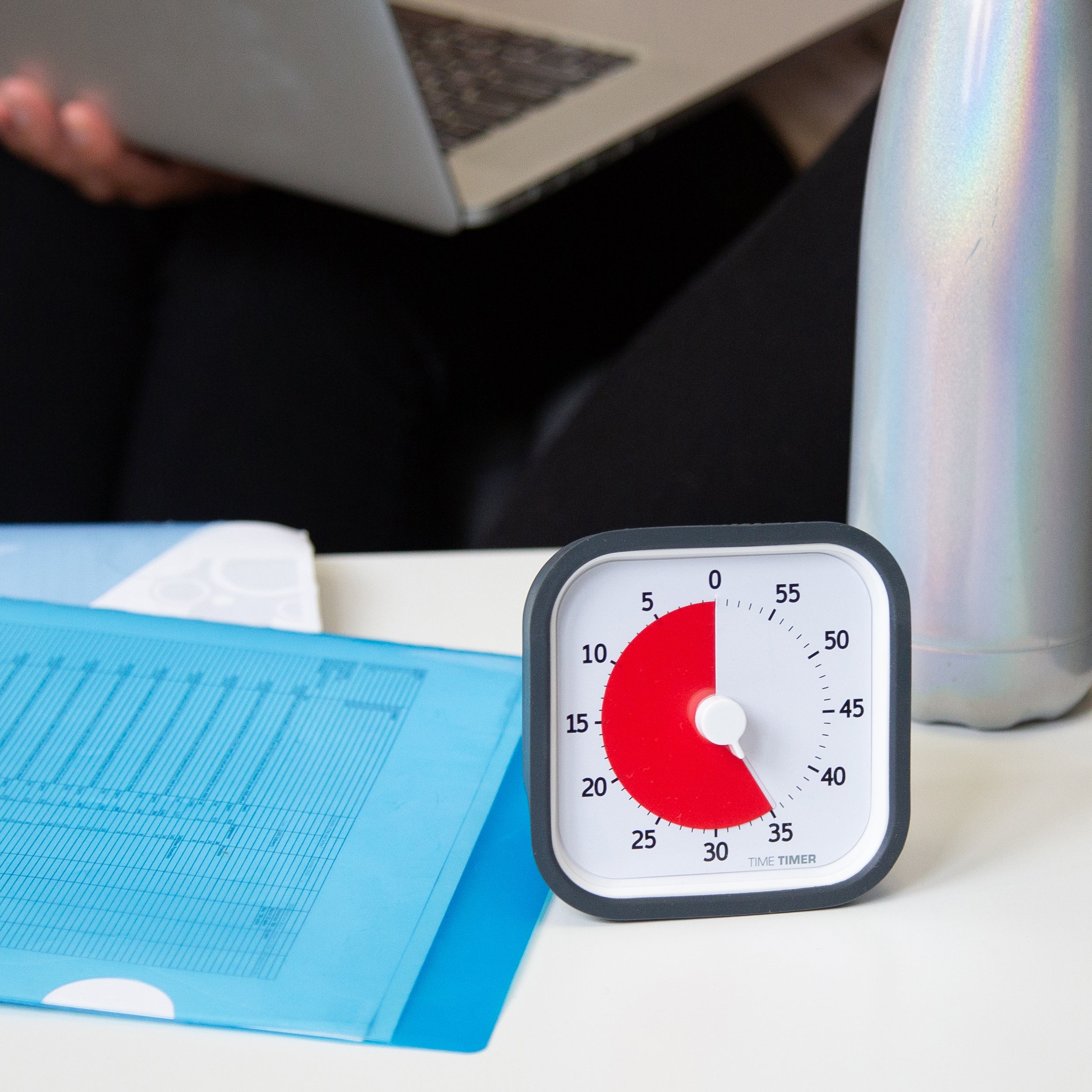 The Time Timer MOD 60 Minute visual timer is shown on a desk with spreadsheets and a water bottle. There is someone in the background on a computer. The red disk is set for 35 minutes.