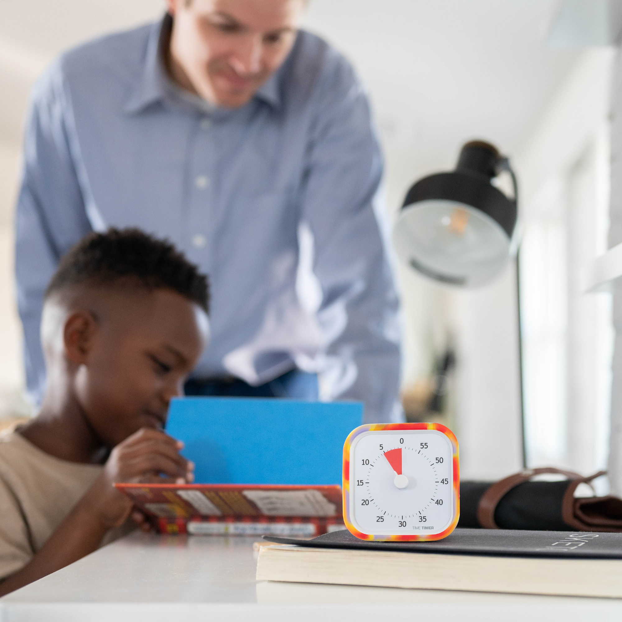 Little boy is reading his book with a male figure standing over him , smiling. A Time Timer is at the front showing how much reading time is left. 